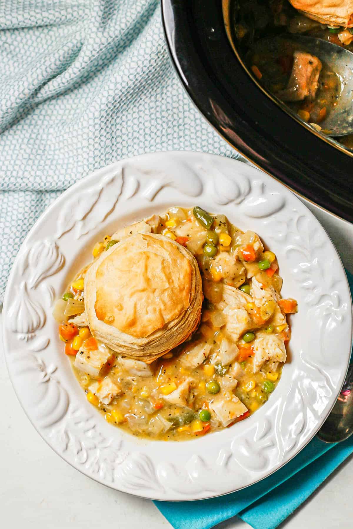 A white soup bowl with chicken pot pie with a biscuit on top beside a slow cooker where it was cooked.