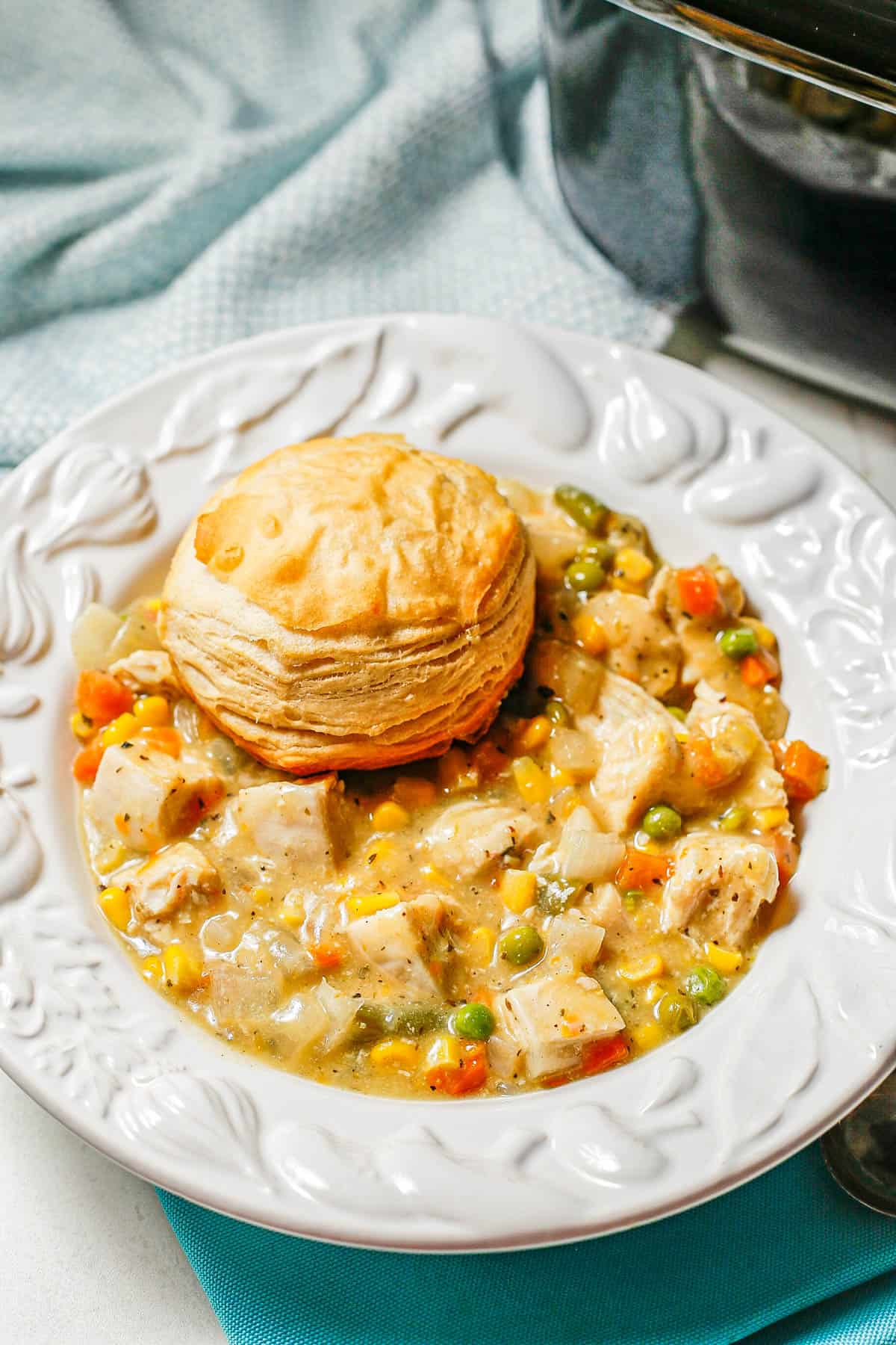 A white decorative soup bowl with chicken pot pie with a biscuit on top beside a crock pot where it was cooked.
