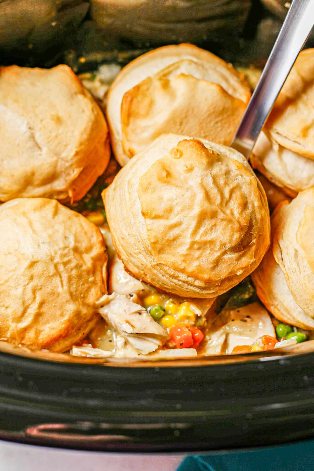 A silver ladle scooping up chicken pot pie and a biscuit from a slow cooker.