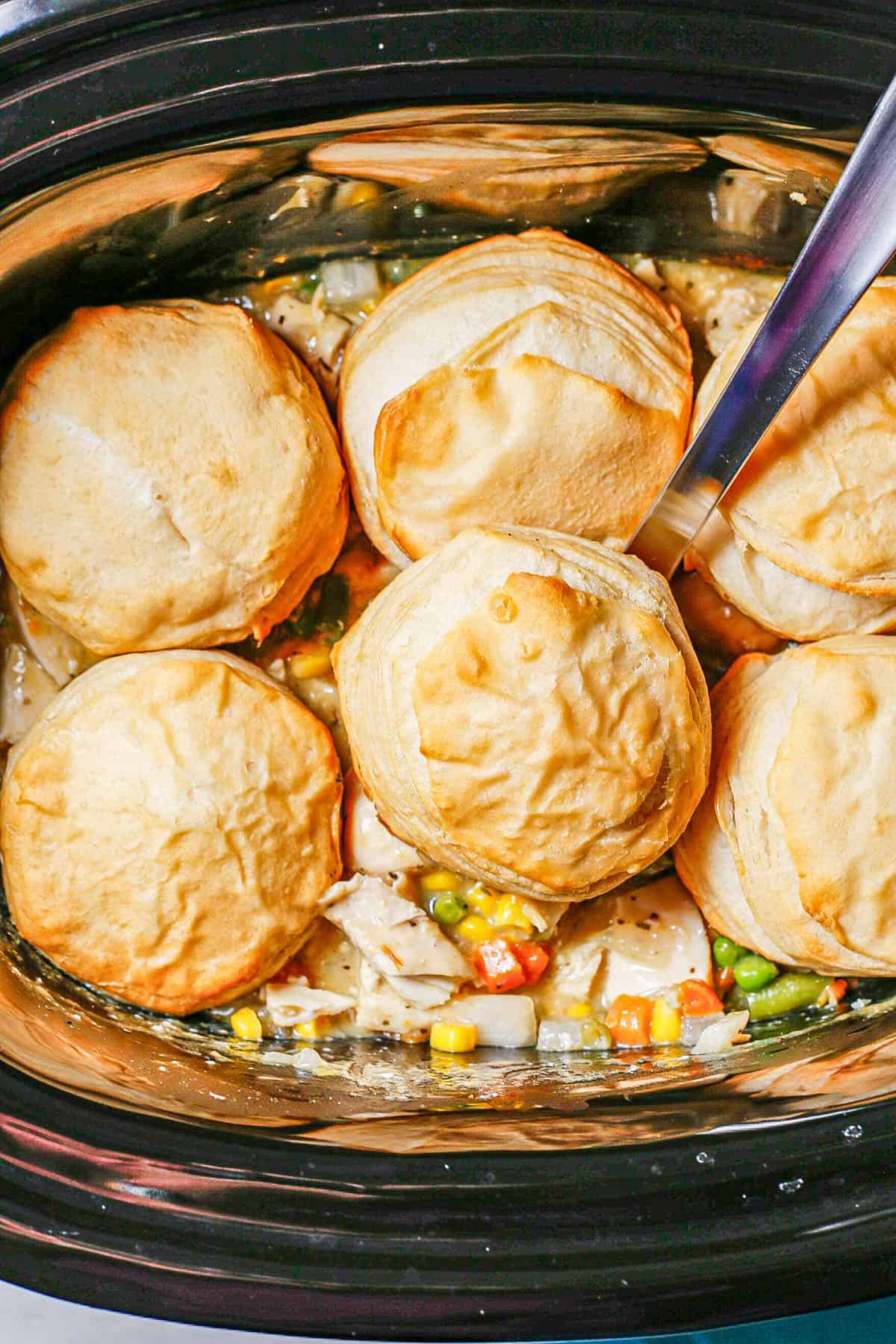 A silver ladle scooping up chicken pot pie and a biscuit from a crock pot.