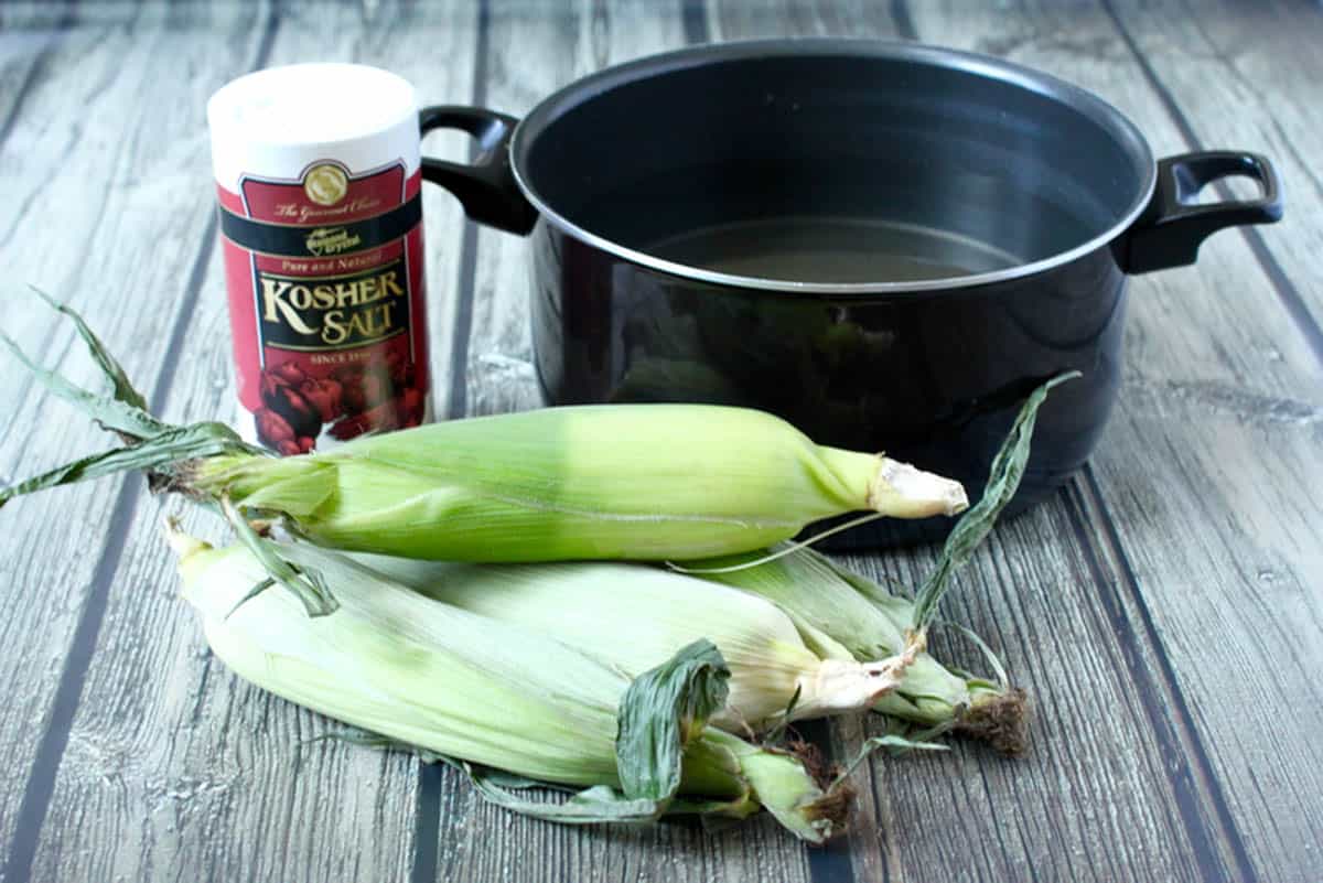 Corn in husks beside a large pot of water and a container of salt.