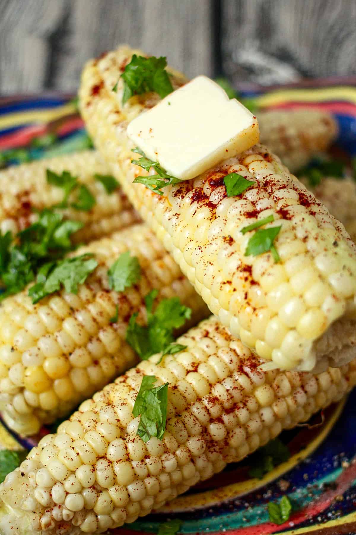 Close up of a corn on the cob stacked on other corn with a pat of butter on top and parsley and chili powdered sprinkled all over it.