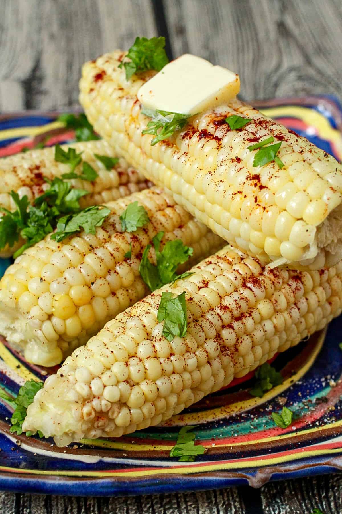 Four ears of corn stacked on a colorful plate with parsley and chili powder sprinkled over them.