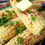 Close up of a corn on the cob stacked on other corn with a pat of butter on top and parsley and chili powdered sprinkled all over it and a text overlay on the photo.