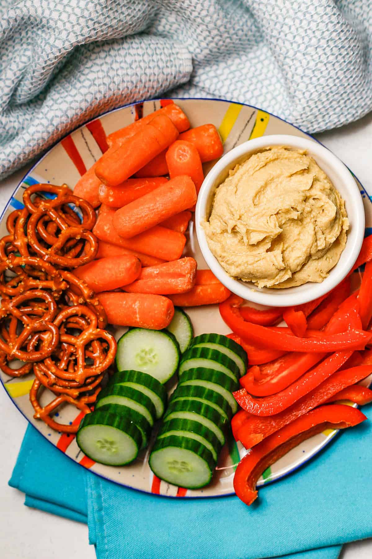 A plate full of fresh veggies and pretzels and a bowl of hummus for dipping.
