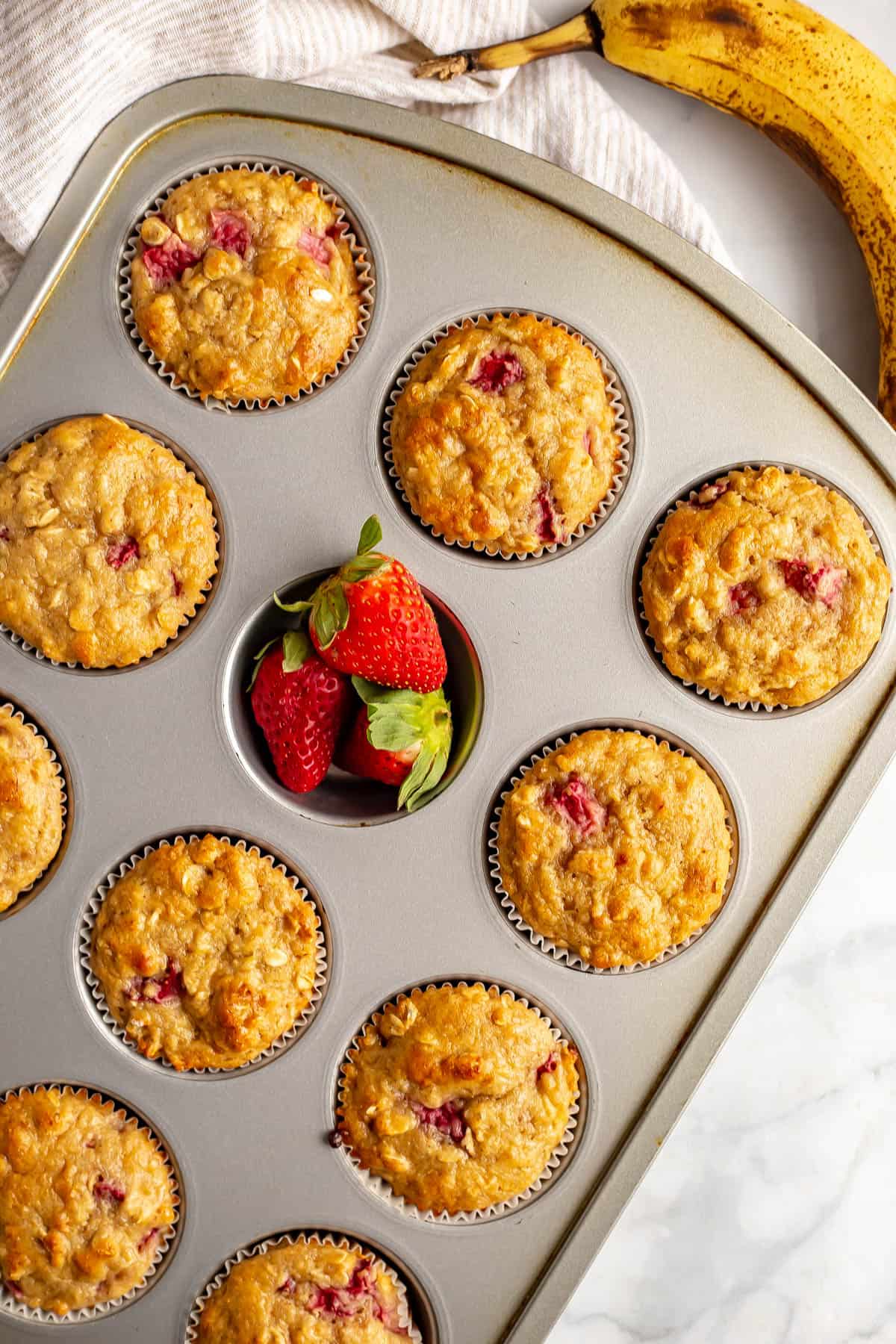 Baked strawberry banana muffins in a muffin tin with one cup filled with fresh strawberries.