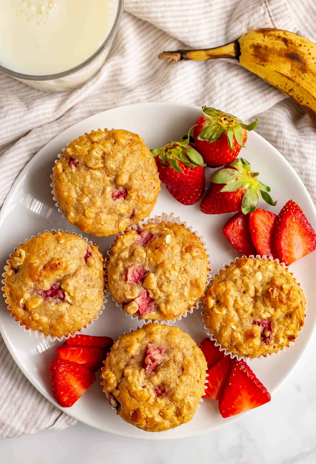 Strawberry banana muffins on a white plate with sliced strawberries on the plate and a glass of milk nearby.