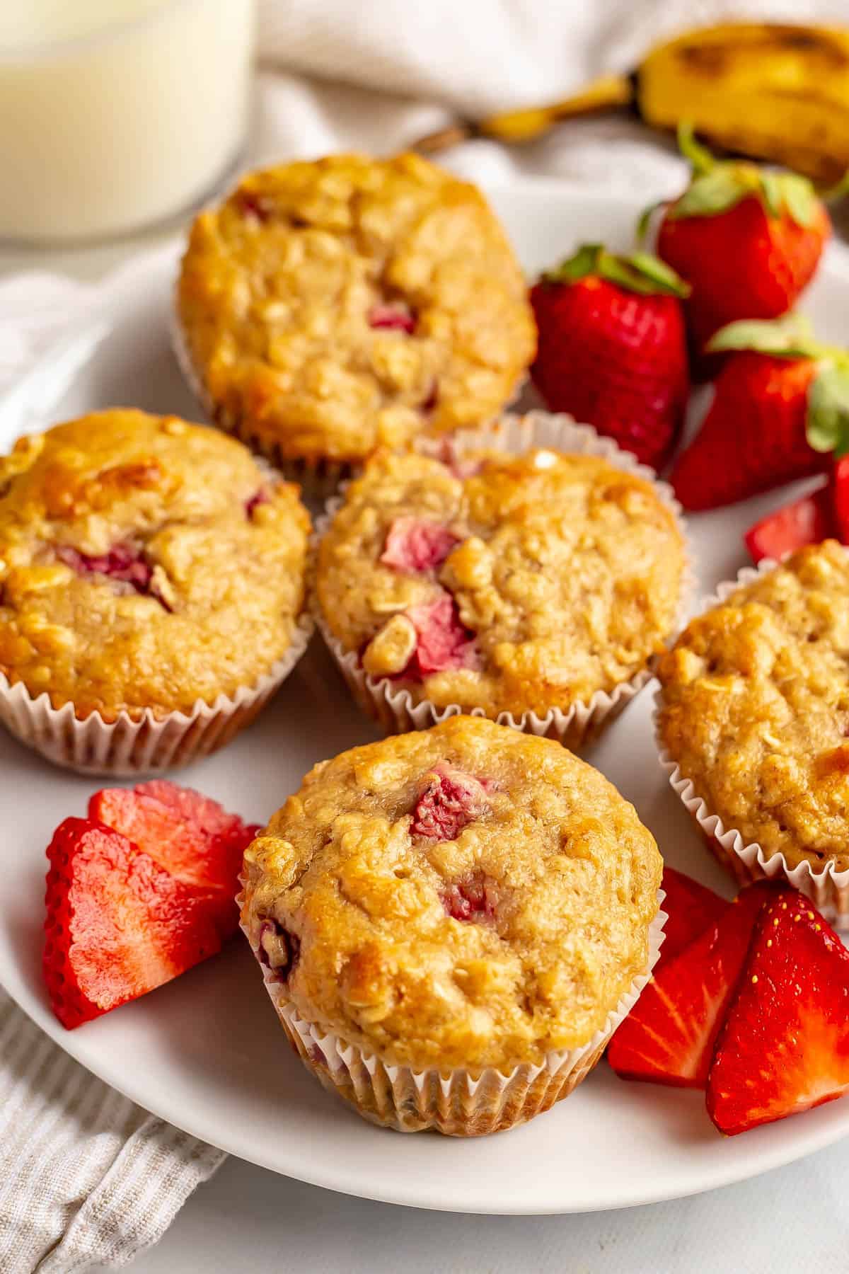 A plate full of strawberry banana muffins along with sliced and whole strawberries with a glass of milk in the background.