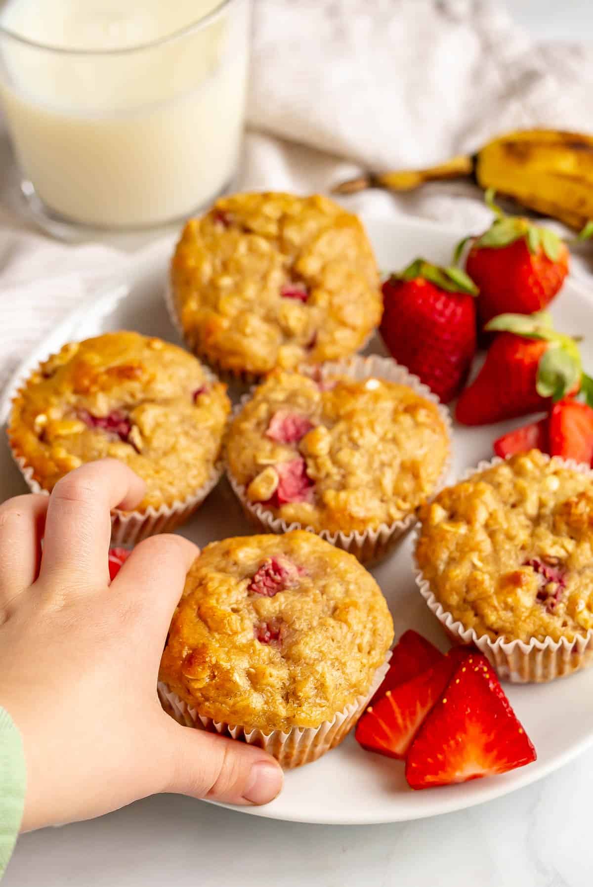 A small hand starting to pick up a strawberry banana muffin from a white plate.