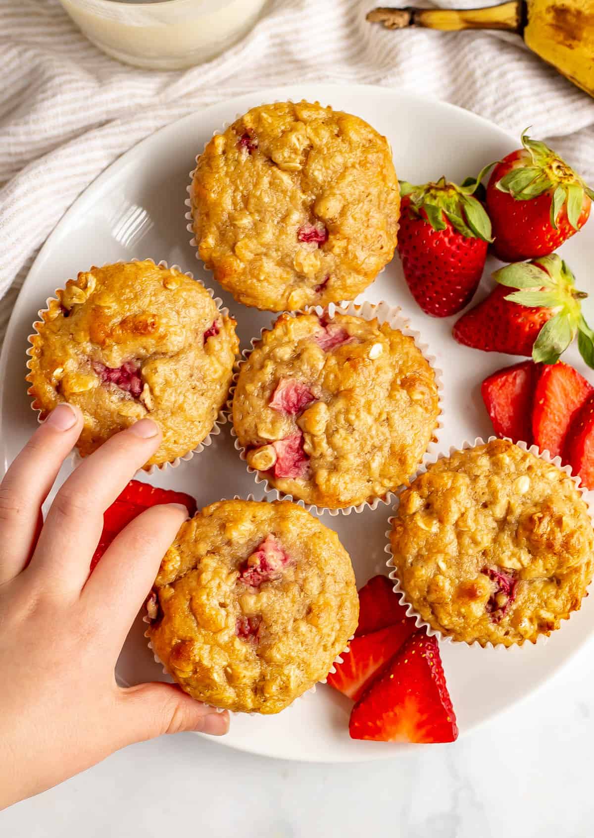 A hand reaching for a strawberry banana muffin on a white plate.