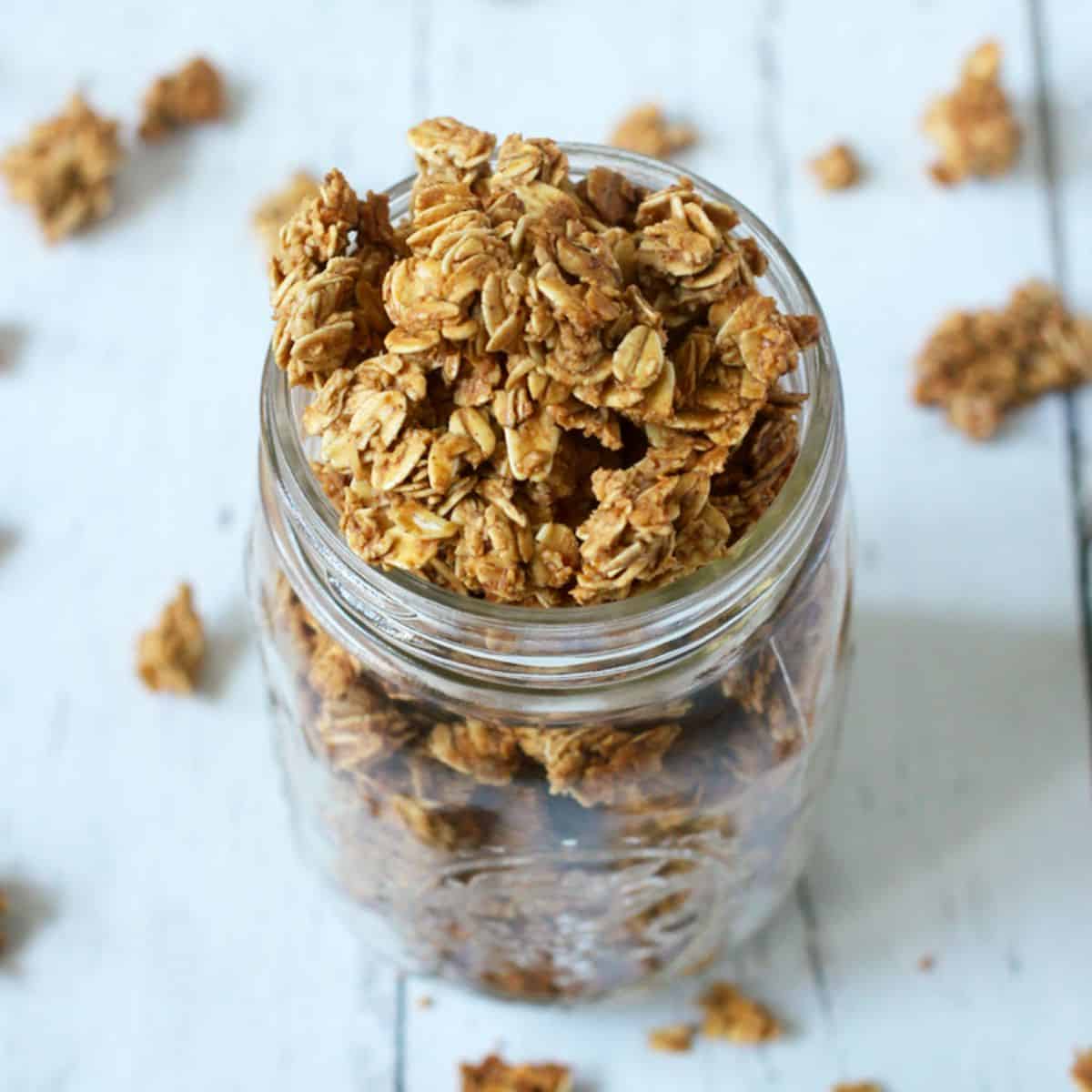 Close up of homemade granola clusters spilling out of a glass jar and onto the counter.