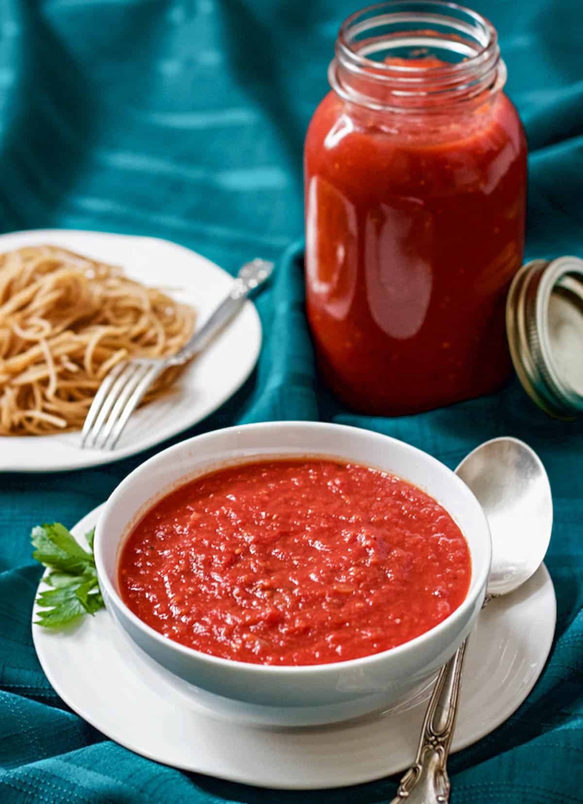 Homemade spaghetti sauce in a white bowl with a jar of extra sauce in the background a bowl of spaghetti noodles to the side.