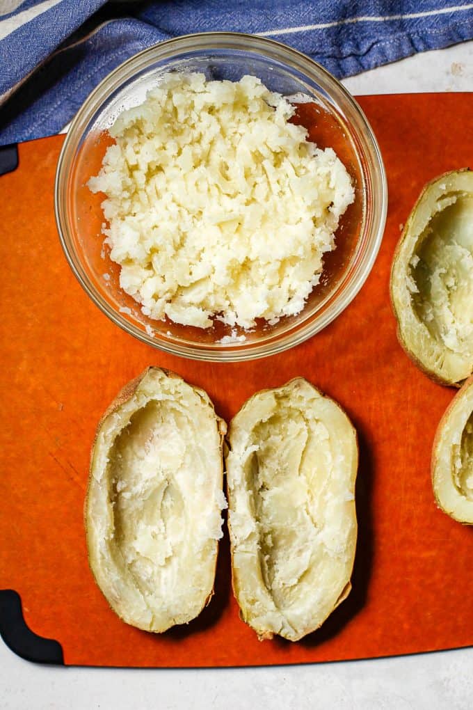 Mashed potato in a bowl with hulled out baked potatoes nearby on the cutting board.