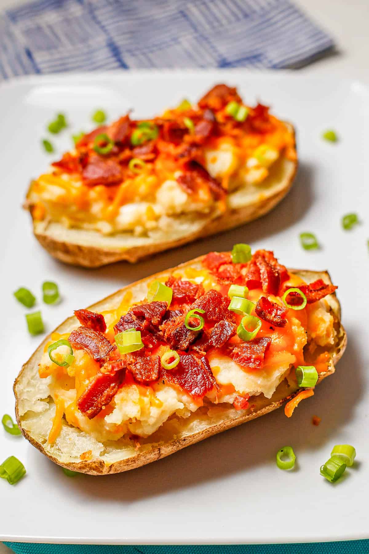 Two halves of a loaded potato with cheese, bacon and green onions on top on a white rectangular serving plate.