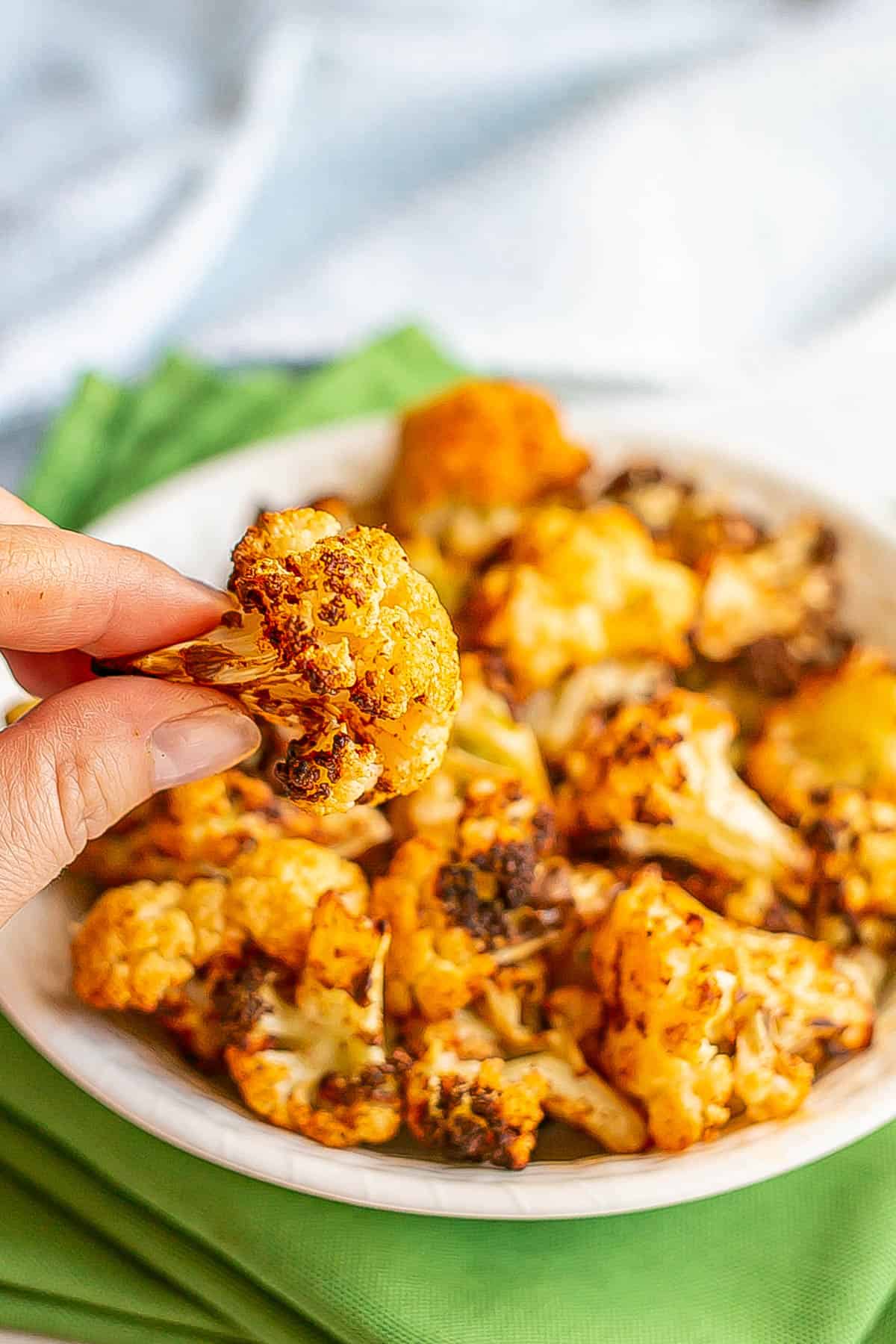 A hand holding up a browned cauliflower floret from a white bowl.