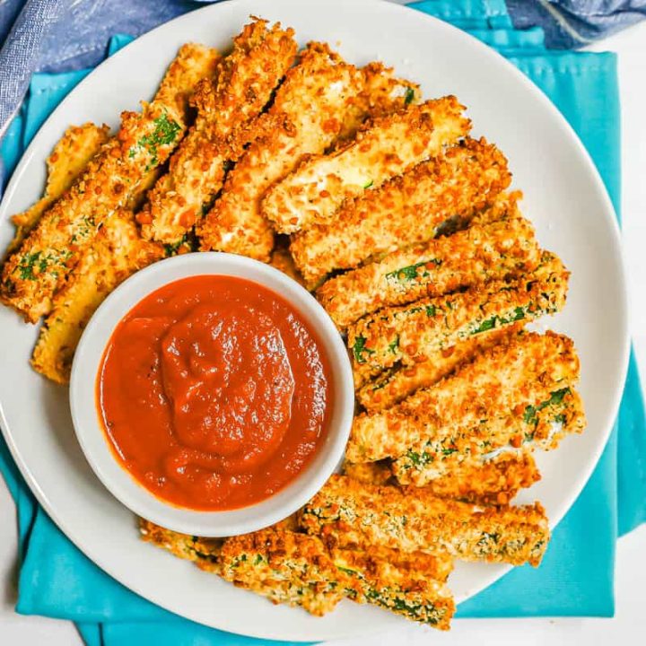 A plate of breaded zucchini fries on a round white plate with a small bowl of marinara.