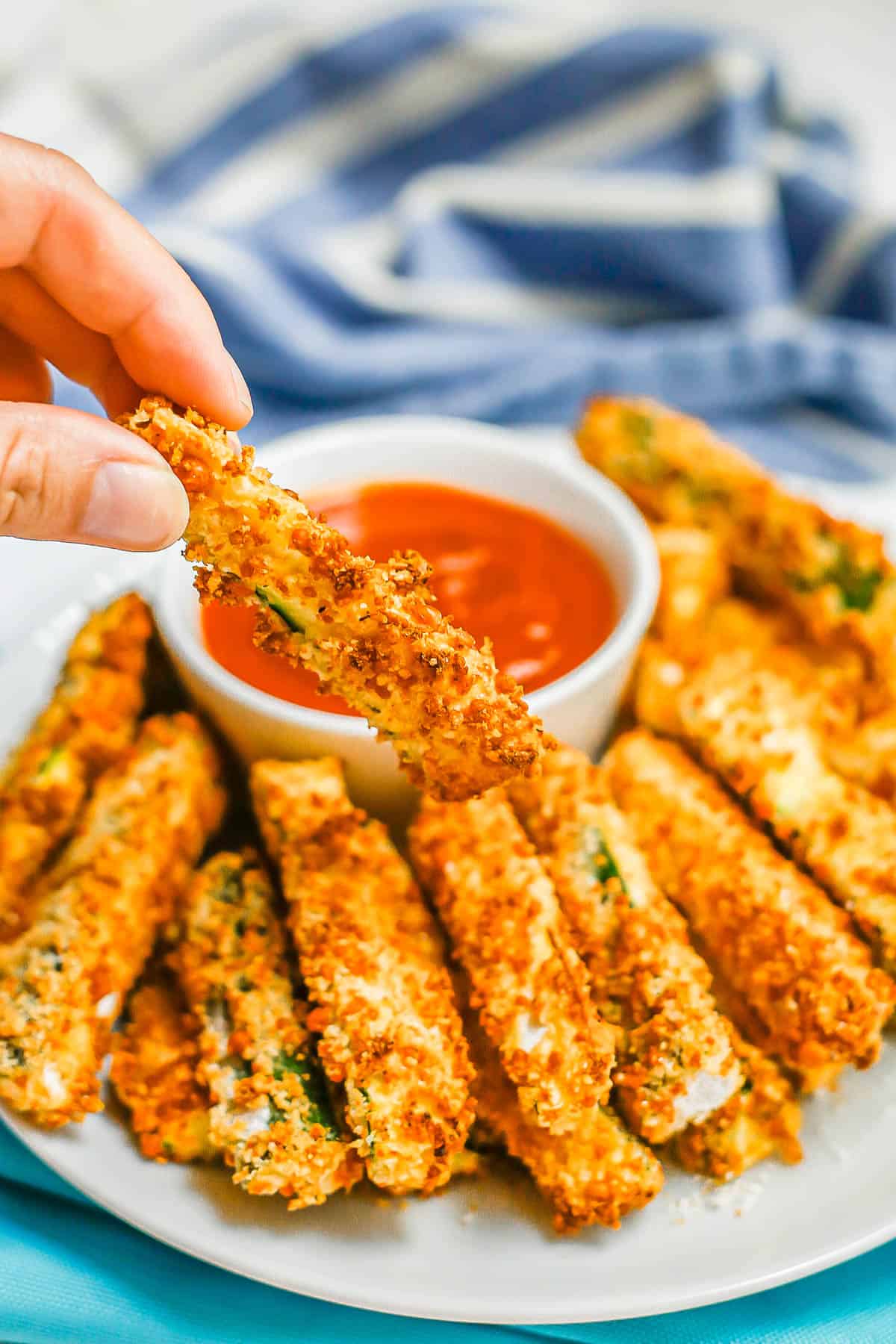 A hand holding up a breaded zucchini stick from a plate with a bowl of marinara for dipping.