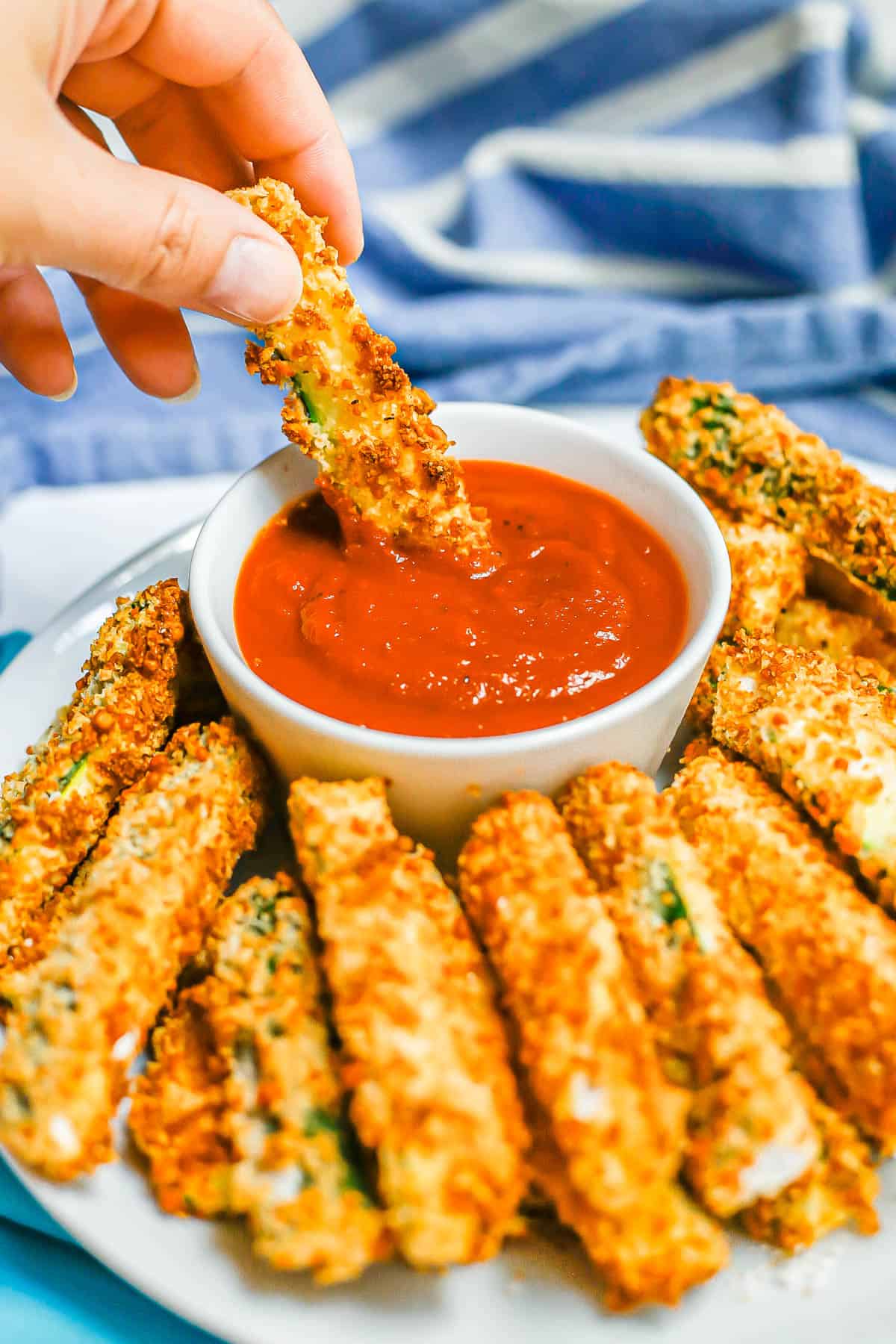 A hand dipping a golden brown breaded zucchini stick in a small bowl of marinara.
