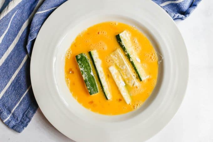 Flour coated zucchini sticks in an egg wash in a low white bowl.