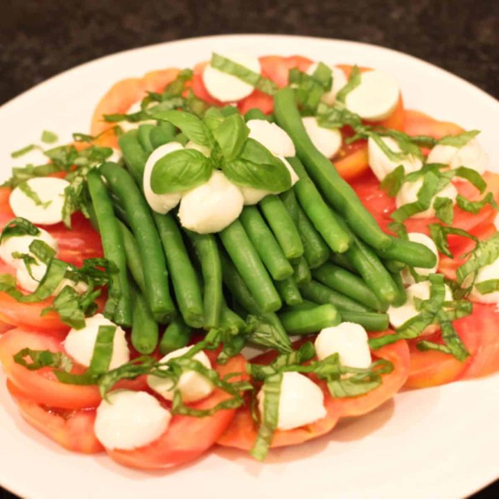 A large white plate with a layer of sliced tomatoes topped with fresh mozzarella and steamed green beans with chopped fresh basil sprinkled on top.
