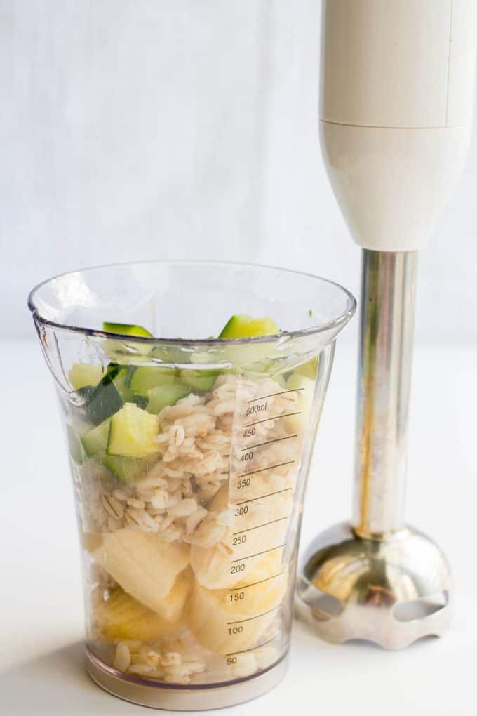 A glass measuring cup with banana, barley and zucchini next to an immersion blender.