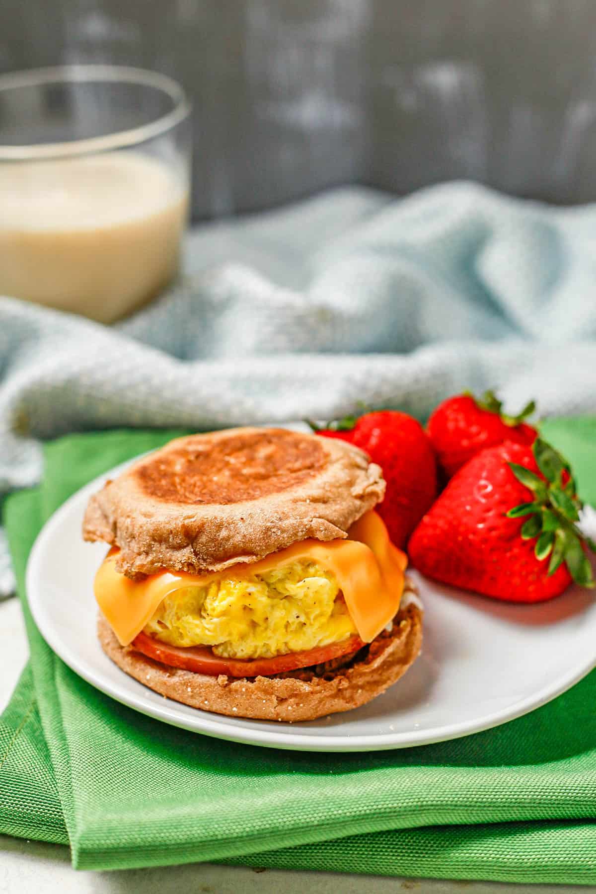 A homemade English muffin sandwich on a white plate alongside some fresh strawberries with a glass of milk in the background.