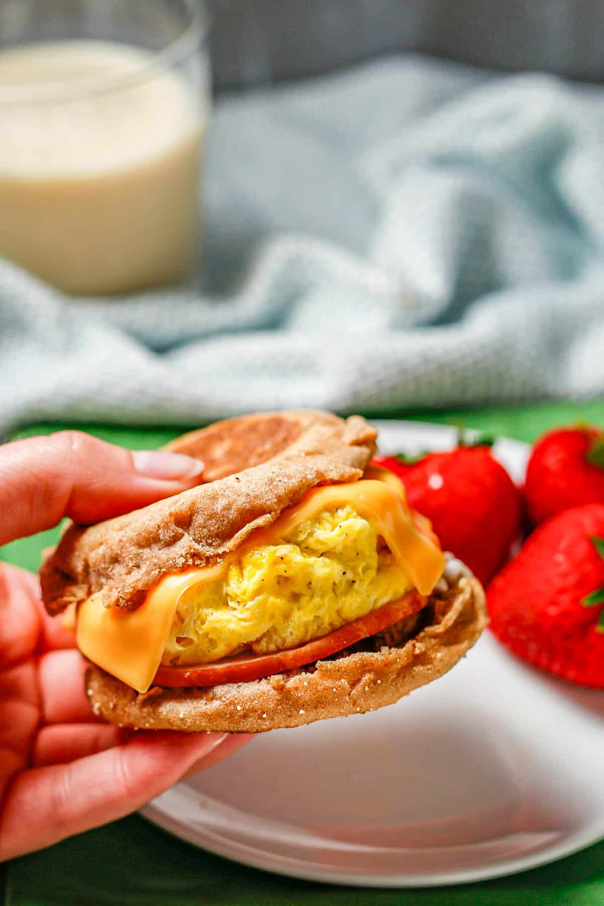 A hand holding up a homemade English muffin sandwich from a white plate with some fresh strawberries on it and a glass of milk in the background.