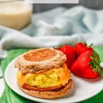 A homemade English muffin sandwich on a white plate alongside some fresh strawberries with a glass of milk in the background and a text overlay on the photo.