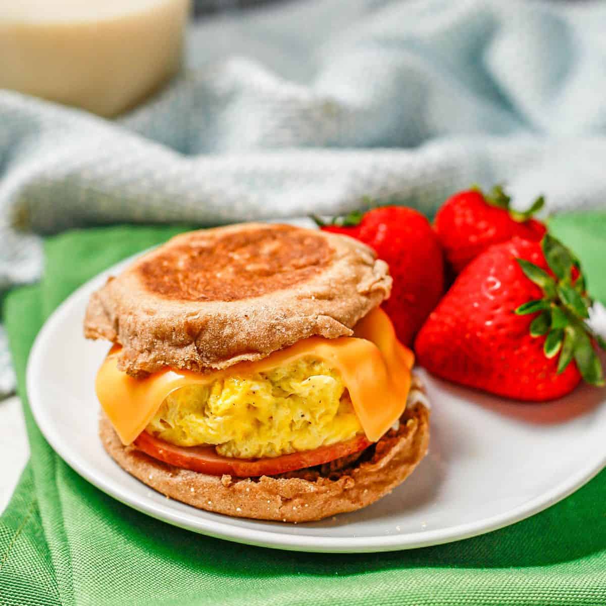 Close up of a homemade English muffin sandwich on a white plate alongside some fresh strawberries with a glass of milk in the background.