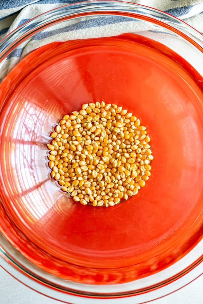 Popcorn kernels in a large glass bowl with a red lid underneath.