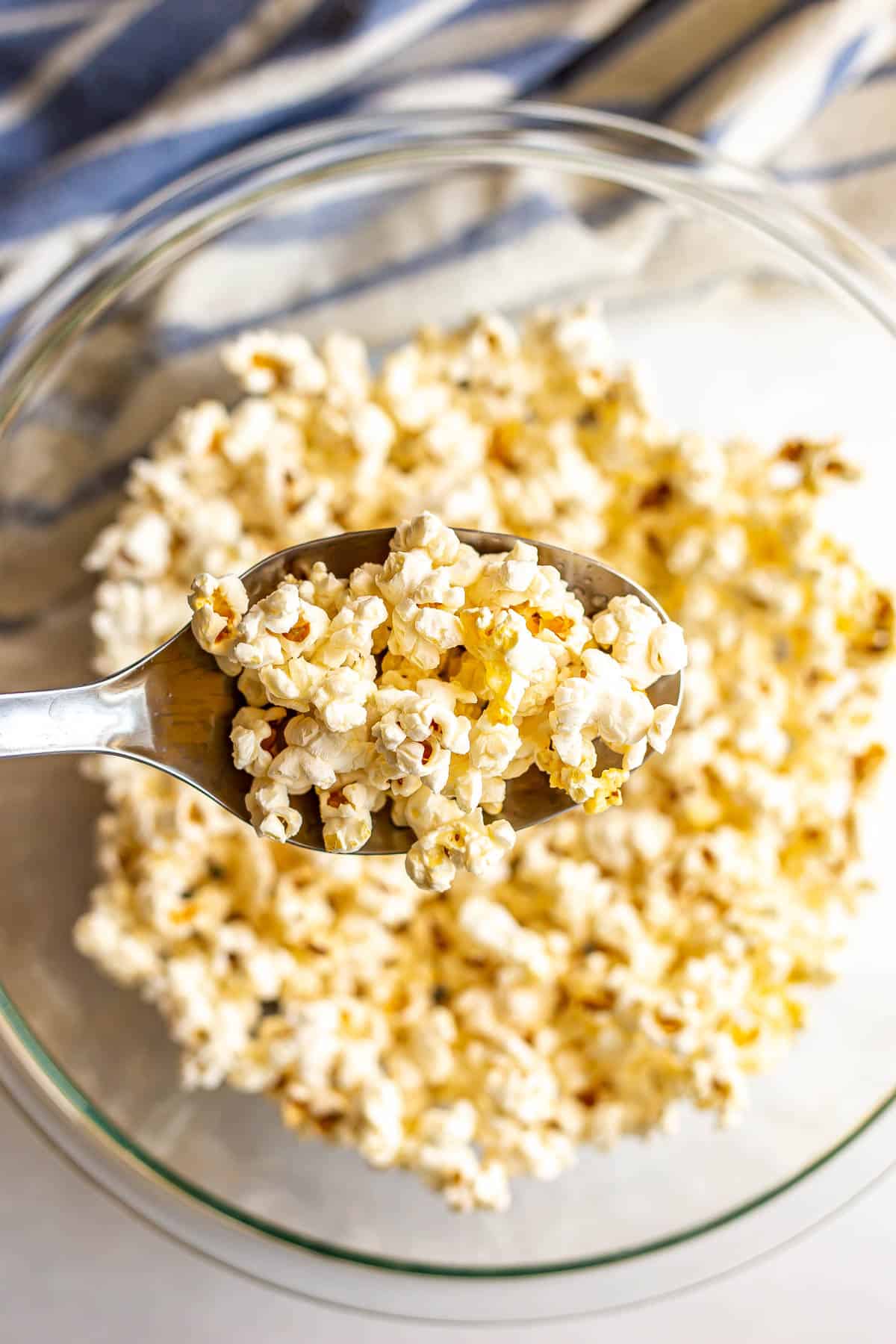 A metal spoon holding up a scoop of popcorn from a large glass bowl.