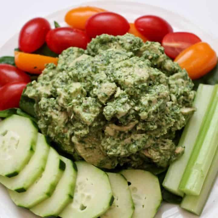 A pile of pesto chicken salad served over baby spinach leaves on a white plate and surrounded by fresh veggies for dipping.