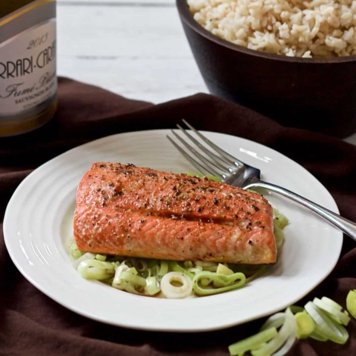 A roasted salmon filet served on a better of tender sliced leeks with a soy sauce mixture drizzled over and a bowl of rice in the background.