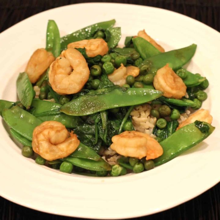 A white bowl with brown rice topped with a shrimp and snow pea stir fry mix with peas and spinach.