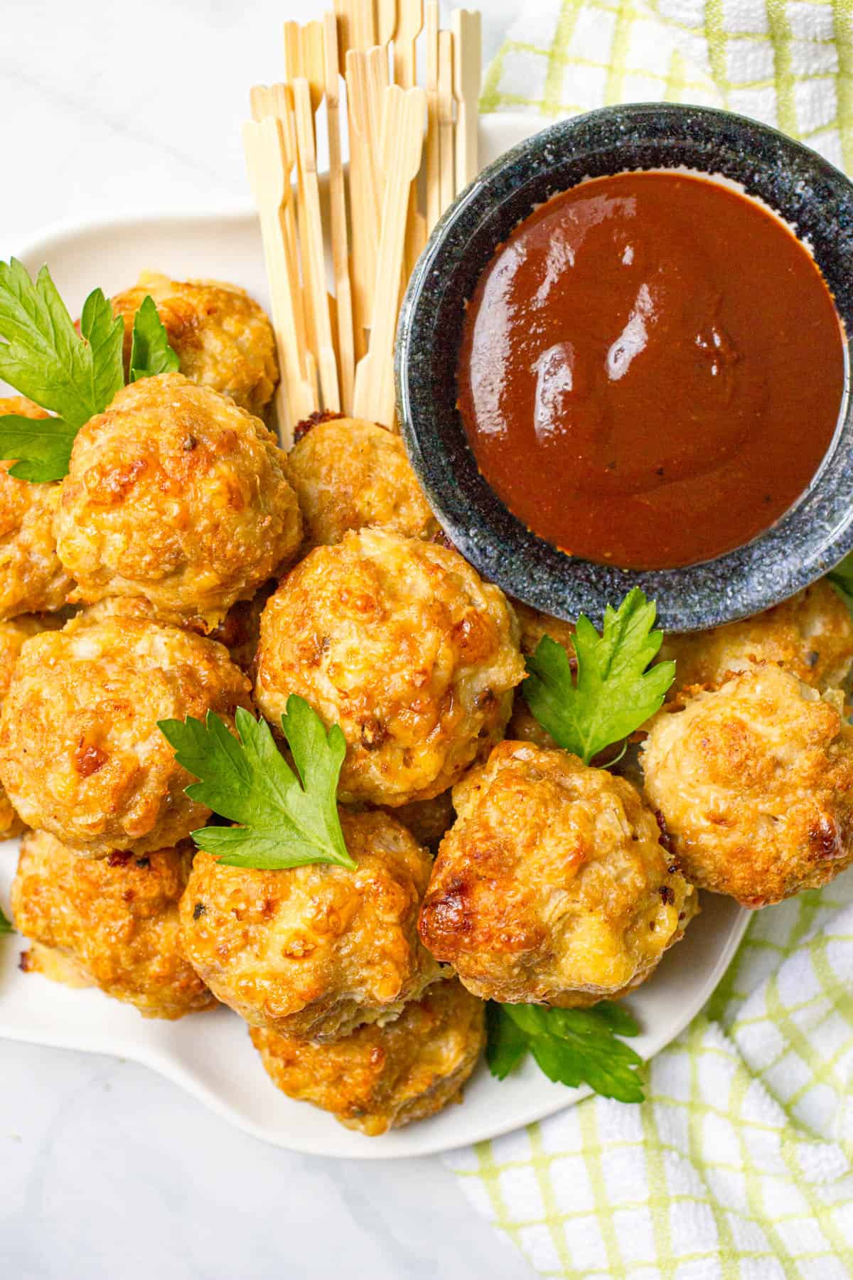 Cheesy chicken meatballs on a white plate with a bowl of BBQ sauce for dipping and leaves of parsley sprinkled over top.