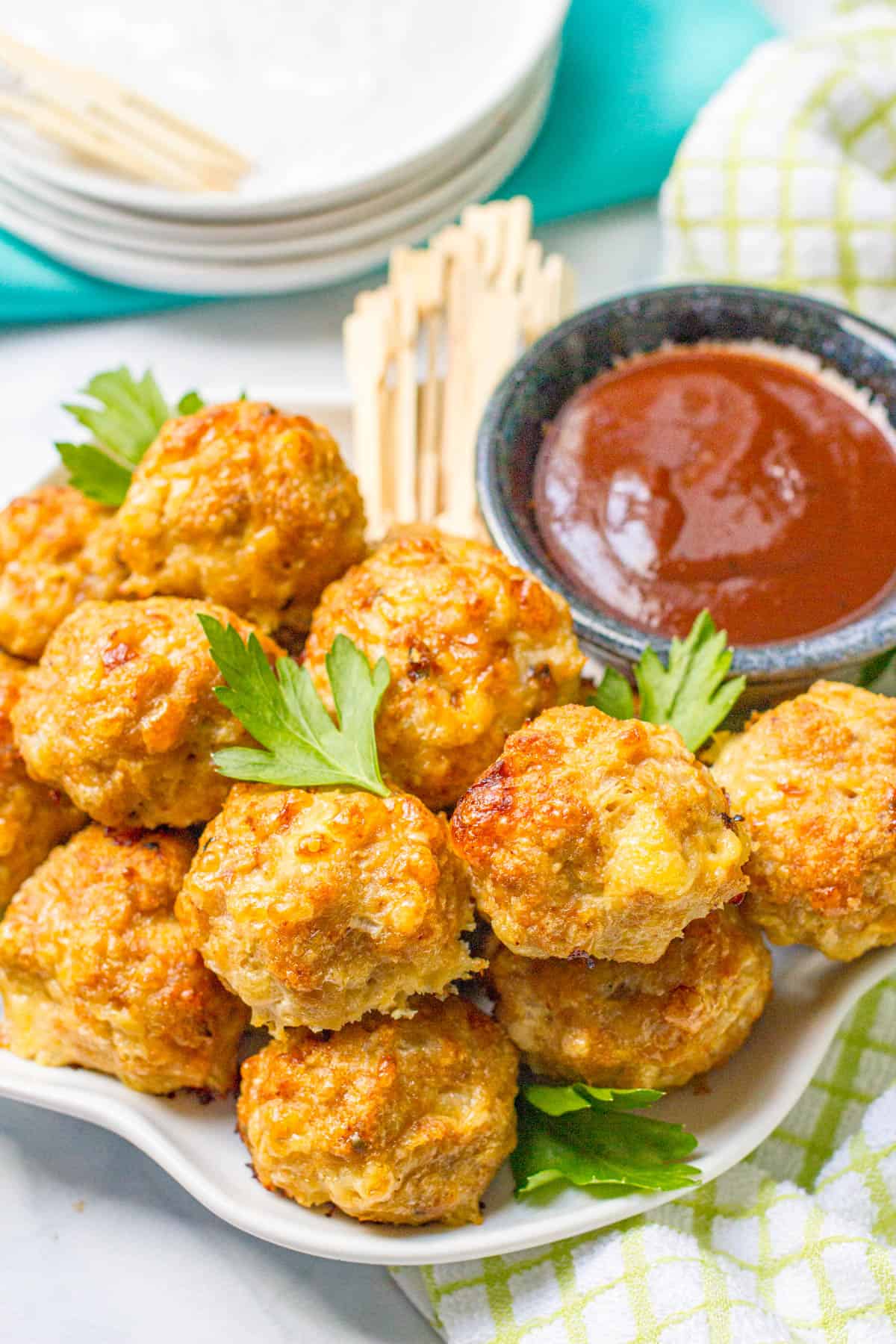 Baked chicken meatballs piled on a white serving plate with toothpicks and a bowl of BBQ sauce in the background.