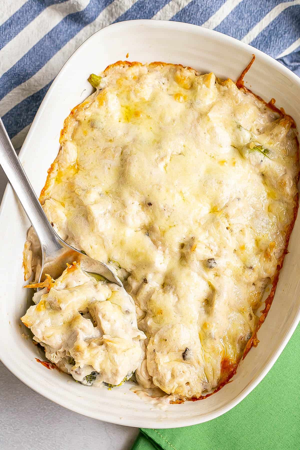 A silver serving spoon resting in a rectangular white casserole dish of baked chicken divan.