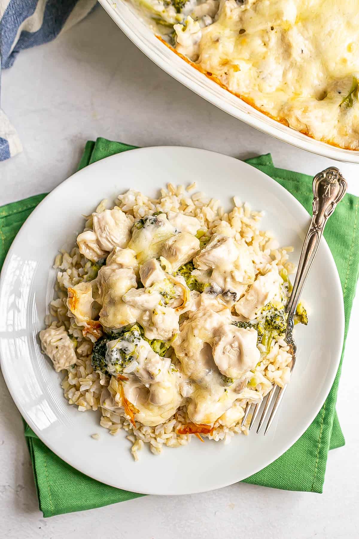 A small white plate set on green napkins with chicken divan served over brown rice with a fork resting on the plate and the baked casserole to the back.