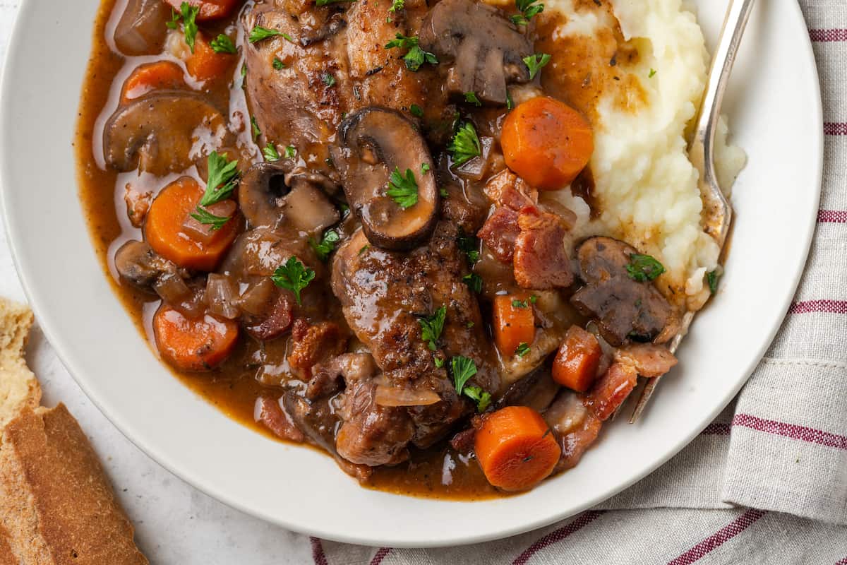 Close up of coq au vin served over mashed potatoes on a white plate with a fork resting on the side.