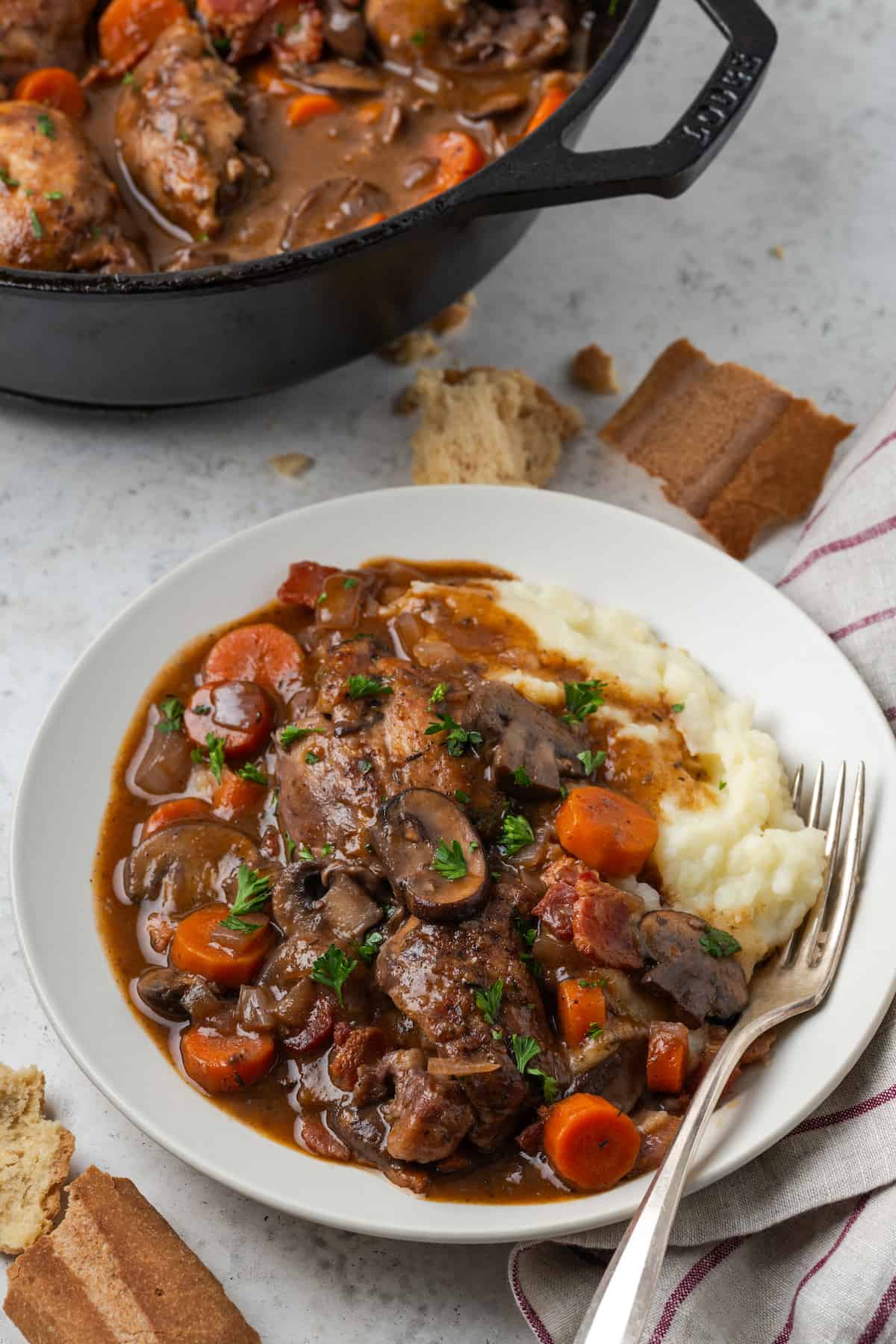 A white plate of coq au vin with mashed potatoes with chunks of bread to the side and a cast iron pot in the background.