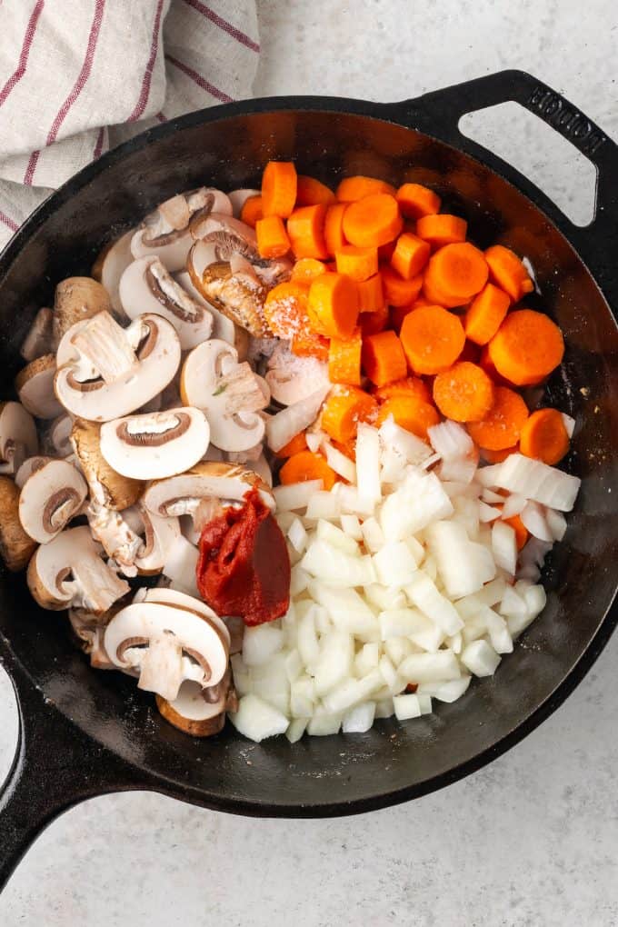 Separate piles of mushrooms, carrots, onion and tomato paste in a cast iron pan before cooking.