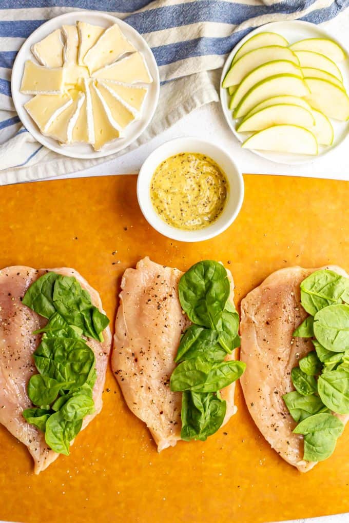 Butterflied chicken breasts on a cutting board with spinach on one side and bowls of brie, apples and mustard to the side.