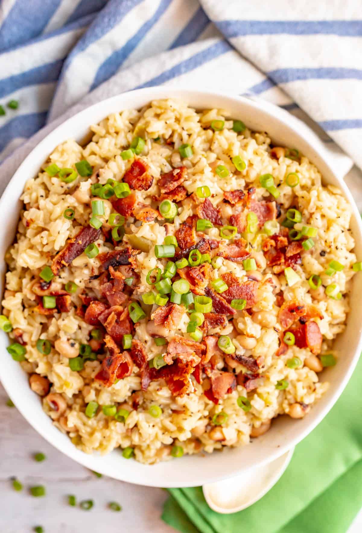 Hoppin John served in a large white bowl with crumbled bacon and green onions on top and green napkins to the side with a blue striped towel in the background.