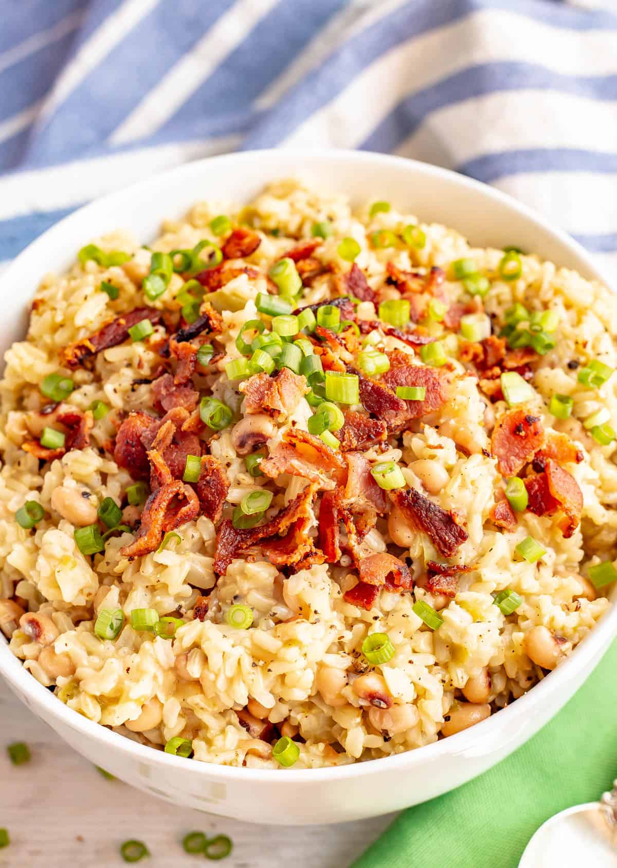 A bowl of hoppin' John topped with bacon and green onions with a spoon resting on a green napkin to the side.