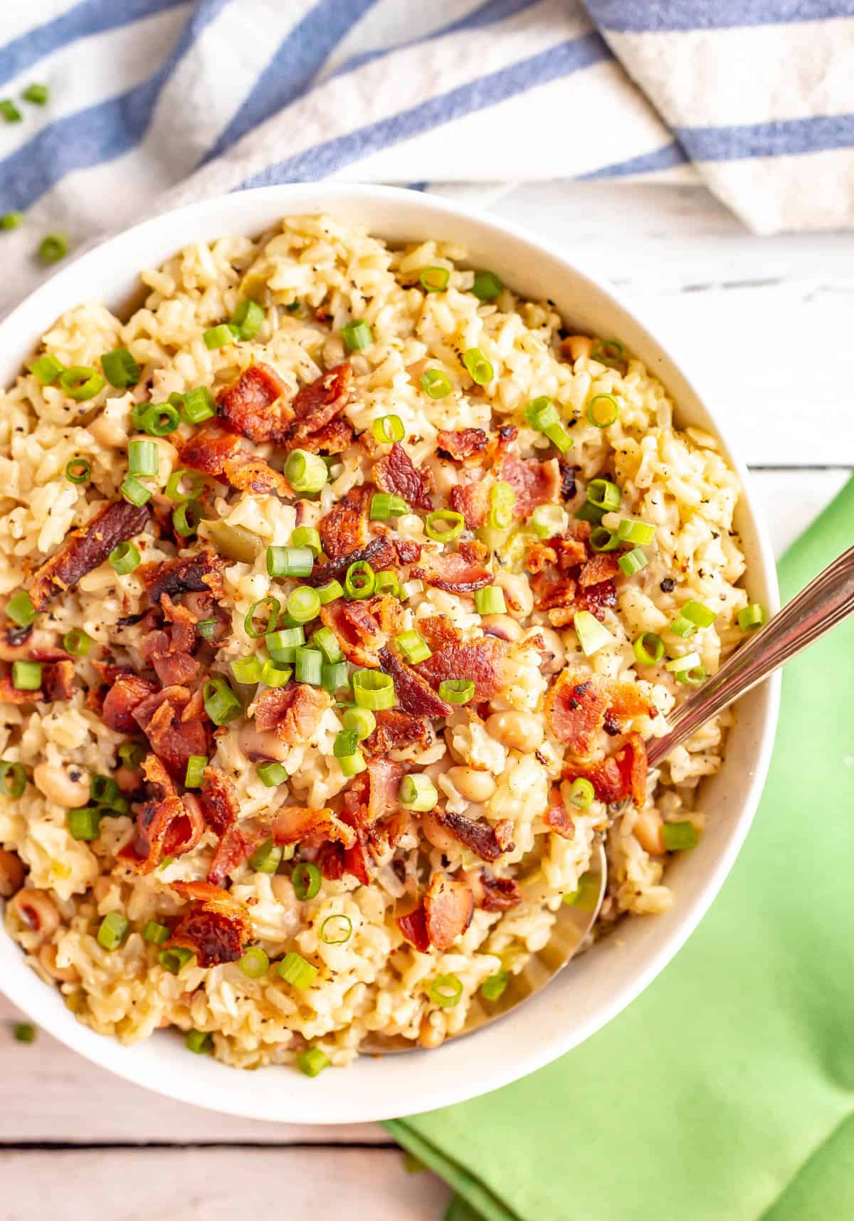 A serving spoon resting in a bowl of hoppin' John topped with cooked, crumbled bacon and green onions.