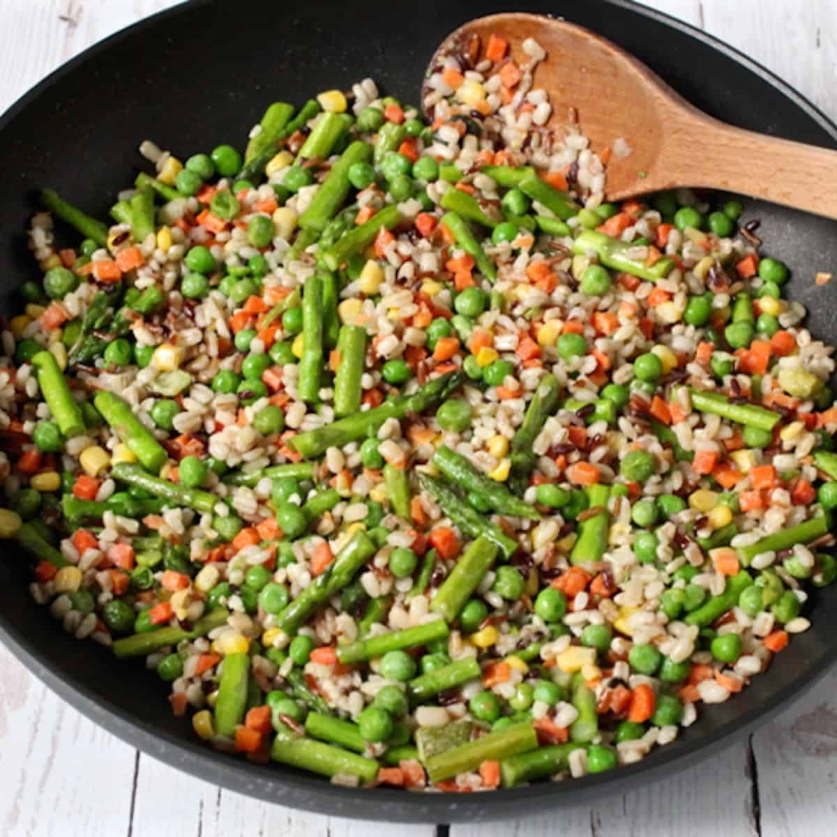 A large skillet with a colorful mixture of grains and vegetables and a wooden spoon resting to the side.