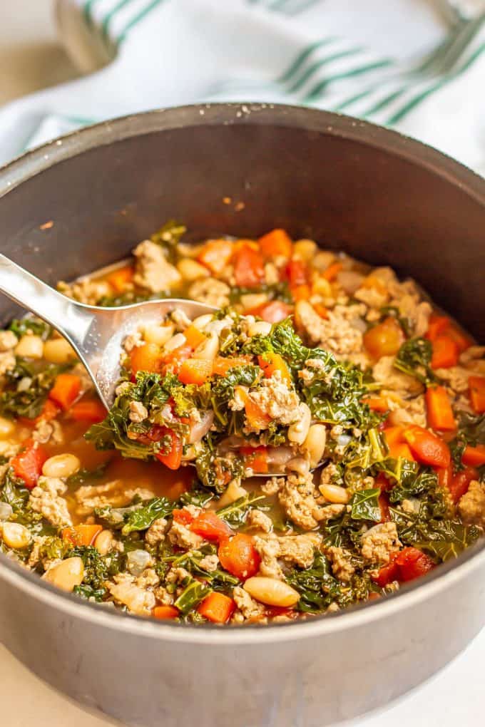 A spoon scooping up a turkey, kale and white bean soup from a large pot.