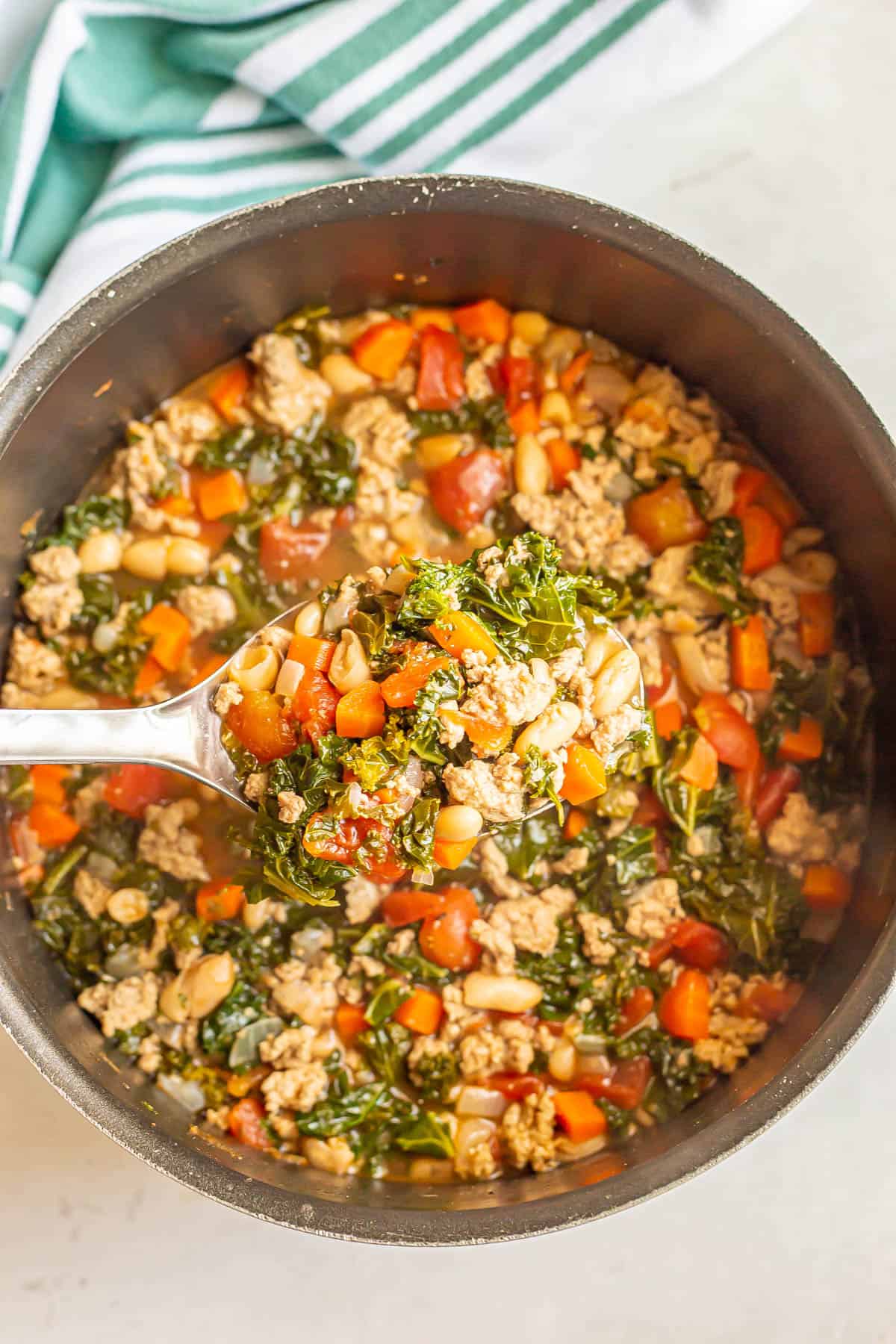 A scoop being held up from a large pot of soup with ground turkey, tomatoes, carrots and kale.