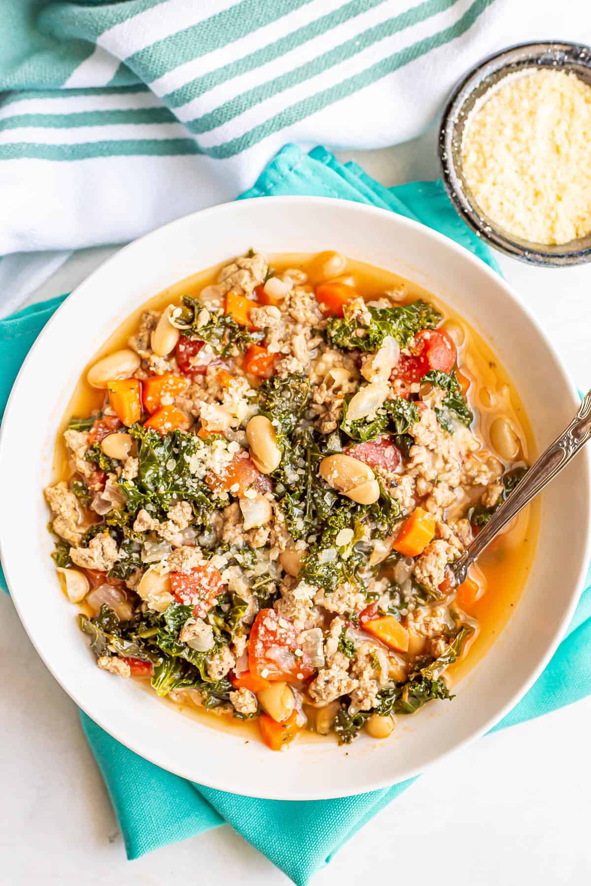 A turkey white bean and kale soup in a white bowl with a spoon resting in it with Parmesan on top and in a bowl to the side.