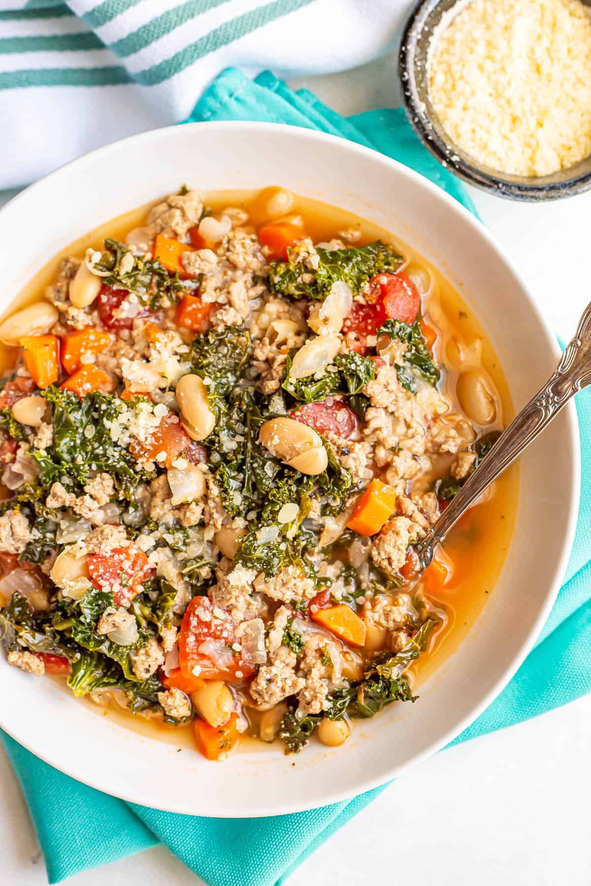 Close up of a turkey white bean and kale soup in a white bowl with a spoon resting in it with Parmesan on top and in a bowl to the side.